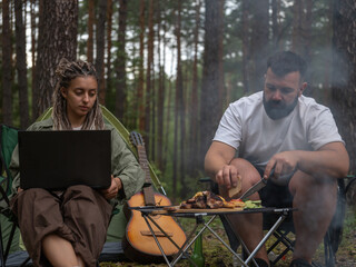 Freelancers enjoying camping trip in nature, combining work and leisure, with man preparing barbecue and woman using laptop near tent and guitar