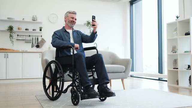 Caucasian man with gray hair sitting in wheelchair using smartphone for video call. Interior setting conveys warmth and modern design, highlighting connectivity, independence, and accessibility.