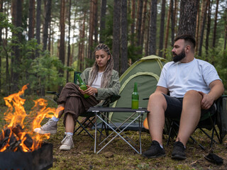 Young couple enjoying a serene camping experience in the forest, relaxing by a crackling campfire, sipping on cold beers, with a cozy tent set against the backdrop of nature