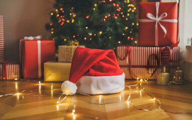 A festive Santa hat on a wooden floor with glowing string lights and wrapped Christmas presents under a decorated tree.