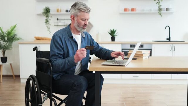 Middle-aged Caucasian man in wheelchair holding bank card, making online payment using laptop in home office setting, expressing independence, accessibility
