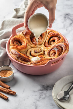 Female hands pouring glaze over freshly baked cinnabon buns in a ceramic baking dish, white marble background. Making cinnamon rolls process
