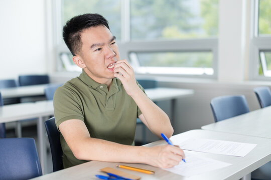 Young man in a bites his nails while solving a test in a brightly lit classroom. He appears anxious or stuck while working through academic problems.