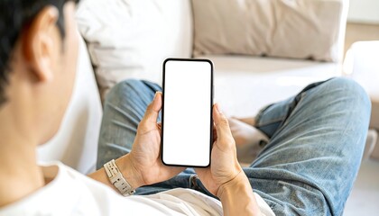 Person on a sofa holds a smartphone with blank screen. Focus is on the device and hands; blurred background features a couch