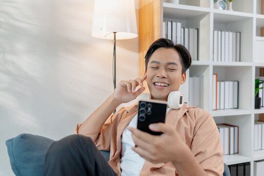 A young man relaxes at home, smiling while using his smartphone. He enjoys leisure time with headphones around his neck, sitting comfortably and feeling casual in a modern living room.