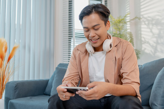 A young man sits on a sofa at home with headphones around his neck, holding a smartphone and playing a mobile game, smiling and celebrating with excitement and happiness. - Powered by Adobe