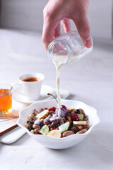 Adult hand pouring creamy milk over a beautiful white bowl filled with fresh fruits, nuts, and healthy ingredients for a wholesome breakfast.