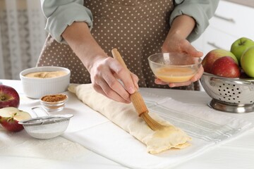 Woman spreading egg yolk onto raw apple strudel at table in kitchen, closeup