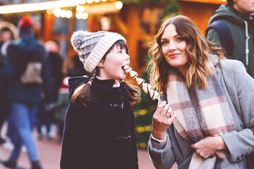 Young mother and daughter eating white chocolate-covered fruits and strawberries on skewers at German Christmas market. Snowy festive fun. Real life, real people, family moment