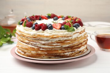 Delicious crepe cake with fresh berries and tea on white wooden table, closeup