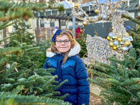 Little cute girl with eyeglasses with lots of xmas trees on Christmas market. Happy child on traditional family market in Germany. Preschooler in colorful winter clothes on sunny day