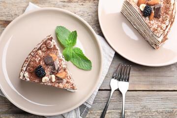 Piece of tasty crepe cake with cocoa powder, chocolate, nuts and blackberries served on wooden table, flat lay