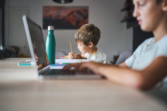 Brothers studying at home together, one writing in notebook with pencil while sibling uses laptop, children focused on learning indoors