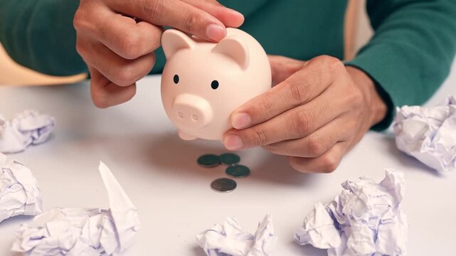 A person puts coins into a piggy bank, but they fall straight through the bottom, symbolizing failed savings, poor financial management, insecurity, and the constant challenge of making ends meet.
