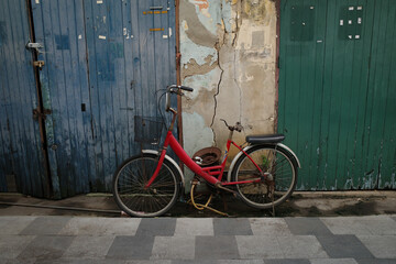 An old red bicycle with a basket stands in front of a cracked and weathered wall with blue and green wooden doors.