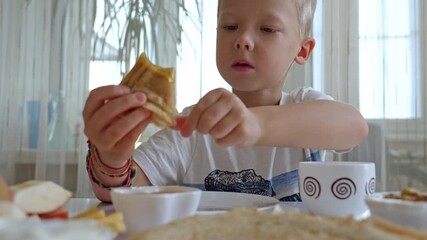 Blond boy dipping golden honey onto crispy waffle, enjoying breakfast with colorful fruits and fluffy pancakes surrounding cheerful meal scene - Powered by Adobe