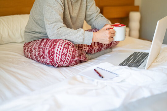 Woman in cozy pajamas sitting on bed, working on laptop while savoring a hot cup of coffee. Notepad and pen nearby, creating a perfect blend of comfort and productivity