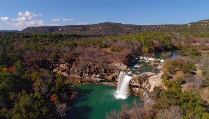 Obraz premium Panoramic view of a waterfall cascading into a turquoise pool, surrounded by autumn foliage and hills