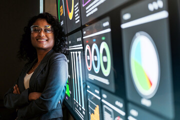 Confident professional standing with folded arms beside large digital dashboard displaying colorful data charts, analytics, and KPIs in a modern office.