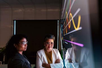 Two professionals examine a large digital screen with colorful data visualizations in a modern office, their reflections visible as they discuss analysis and strategy.