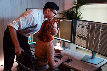 Two colleagues work together at a dual-monitor desk in a modern office, reviewing code or data at night. One stands and guides while the other types, screens glowing in low light.