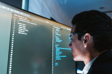 Software developer wearing glasses focuses on code displayed on a large monitor in a modern office, reviewing and debugging source files on a wide screen workstation.