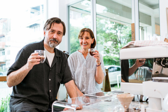 Smiling adults enjoying espresso in a bright modern cafe beside a professional coffee machine, raising cups at the counter with natural light and urban view through large windows. - Powered by Adobe