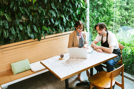 Two colleagues in a cafe with a laptop discuss work beside a lush indoor green wall, suggesting consulting or small business planning in a modern, casual workspace.