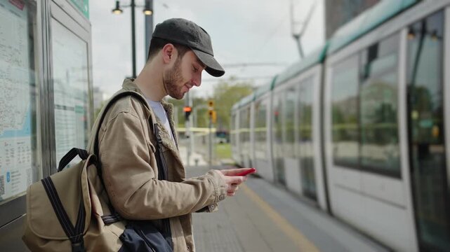 Young man commuter using phone and waiting for public transport at tram train station in the city