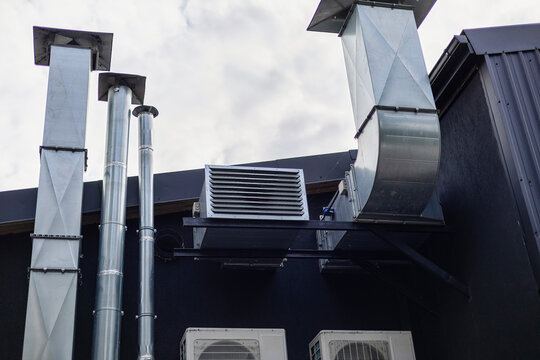A commercial building features a complex ventilation system with silver ducts and exhaust pipes positioned prominently on the rooftop against a cloudy sky.
