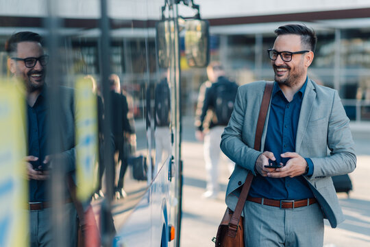 Smiling businessman using smartphone while walking near bus at airport or bus station - Powered by Adobe
