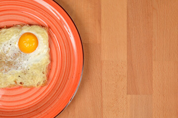A top-down shot of a cheese and fried egg toast on an orange plate, positioned on the left side of a wooden surface, leaving copy space