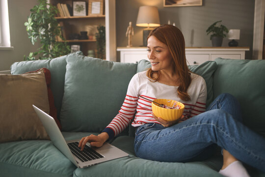 A person with long red hair sits comfortably on a couch, using a laptop while enjoying a bowl of snacks in a cozy living room setting filled with plants and warm light.