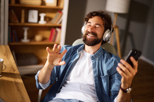 A man with a beard wears headphones and smiles while holding a smartphone. He is seated in a cozy home office, enjoying his music in a relaxed atmosphere filled with books.