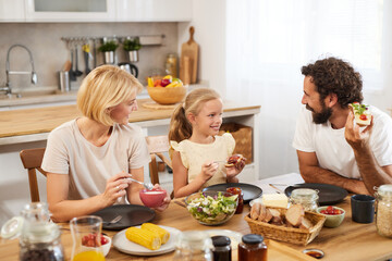 In a bright kitchen, a family gathers around a wooden table for breakfast. Parents and a young girl...