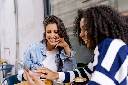 Happy young woman with friend using mobile phone at sidewalk cafe