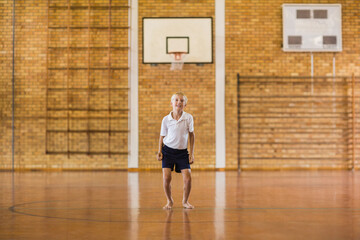 Barefoot boy wearing white polo and shorts standing in gym with basketball hoop and climbing frame