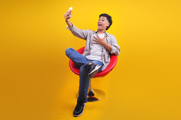 Full Body Of Surprised Young Asian Male Sitting On Chair While Using Smartphone