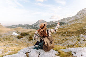 A young female hiker with a backpack and hat sits on a rock, looking at the mountain scenery. A woman on a hiking trail feels free and enjoys the adventure. Concept of travel, freedom.