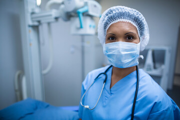Female doctor standing in radiology suite wearing blue scrubs by X-ray machine with surgical mask