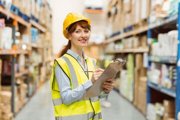 Female warehouse inspector wearing vest with clipboard inspecting inventory in warehouse aisle