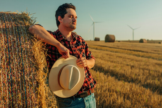 Stylish cowboy in flannel shirt holding hat in wheat field at sunset