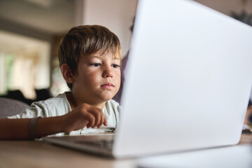 Young boy concentrating on laptop while studying online at home indoors