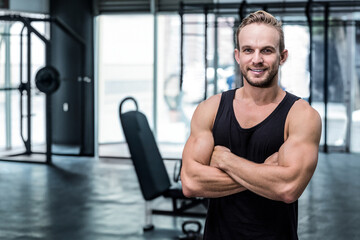 Muscular man in tank top standing with arms crossed at gym near weight machines, copy space