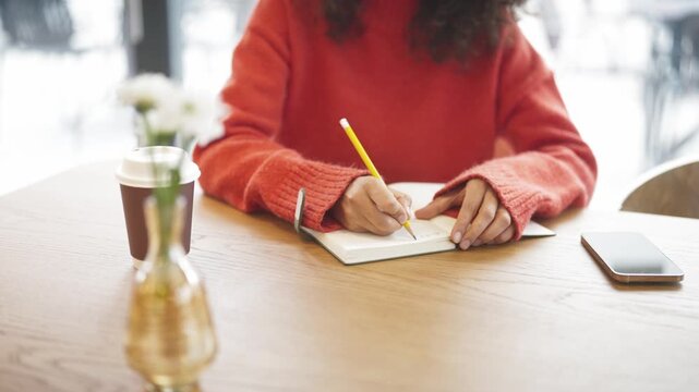 Close up of woman in red sweater writing notes with pencil in notebook at cafe table, takeaway coffee cup nearby. Female with curly hair studying or journaling indoor, smartphone placed on wooden desk