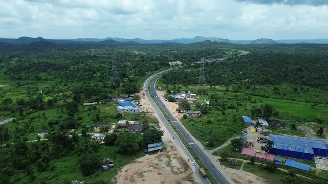 Aerial view of the stark grey highway cutting through the lush green landscape with buildings on either side, Bundu, Jharkhand, India.
