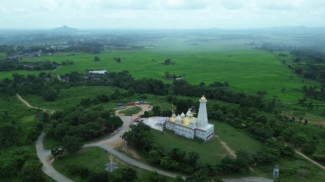 Aerial view of a Surya Mandir temple with white walls and golden domes, nestled among lush green trees and open green fields, Bundu, Jharkhand, India.