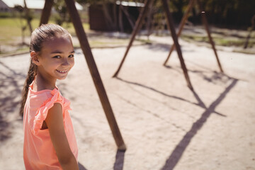 School-age girl standing on sandy playground at suburban park with wooden swing frames, copy space