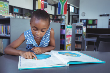 African American school-age girl leaning over open atlas in public library while pointing at map
