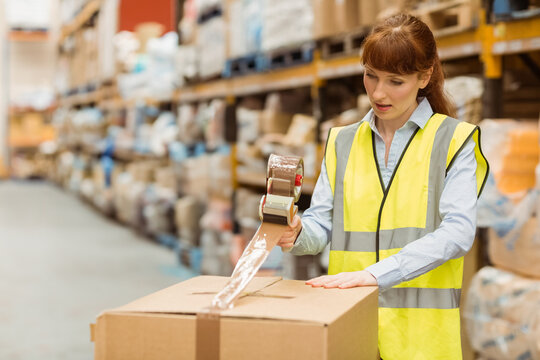 Female worker in hi-vis vest sealing cardboard box with tape dispenser in storage aisle, copy space - Powered by Adobe